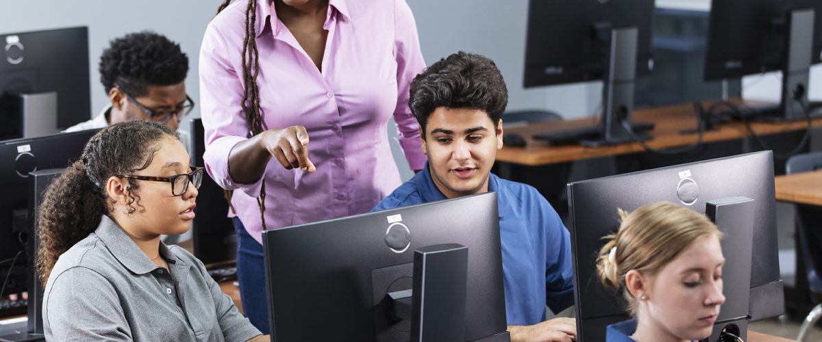 Five multiracial high school students in a computer lab, each using a desktop PC. The teacher, a mature African-American woman, is helping a student, talking and pointing to her monitor.
