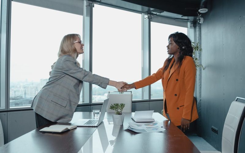 Two businesswomen shaking hands in a modern office setting, symbolizing partnership and agreement.