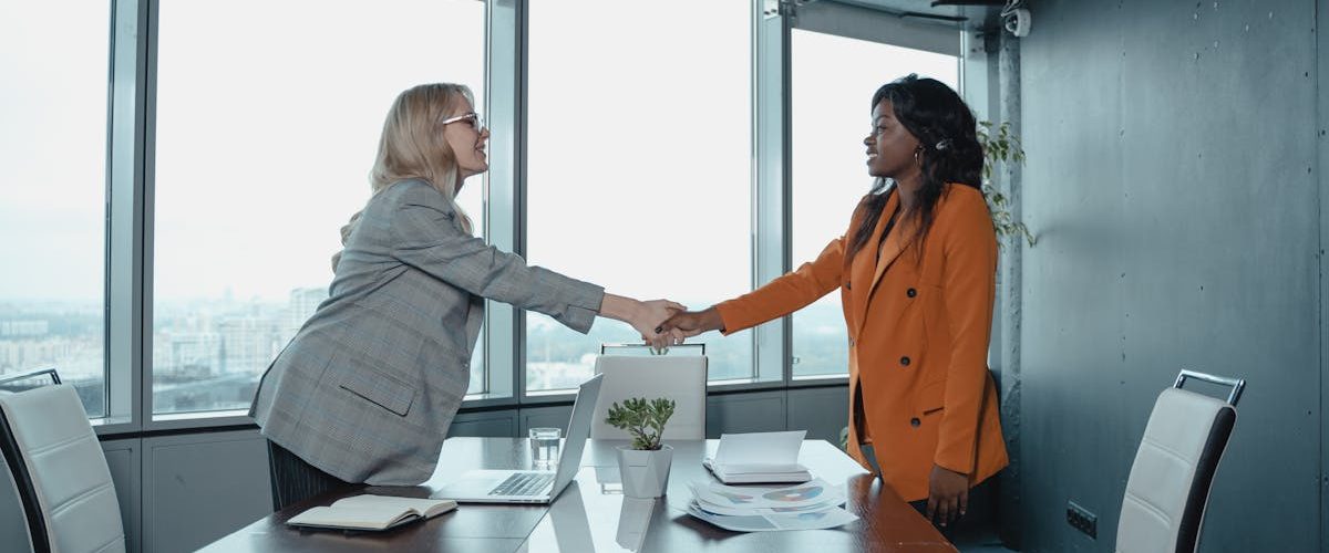 Two businesswomen shaking hands in a modern office setting, symbolizing partnership and agreement.