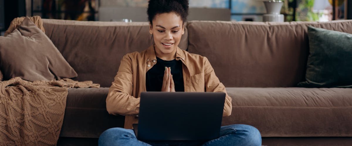 A smiling young woman sits cross-legged on a pouf, using her laptop in a cozy home setting.
